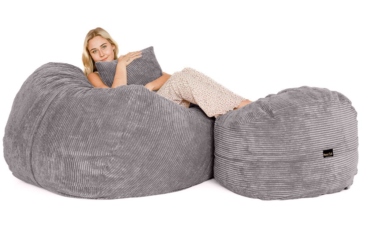 A person sitting on a large gray corduroy bean bag chair with a footrest beside them, reading a book.