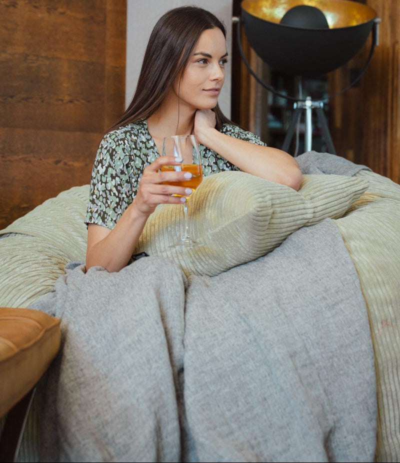 Woman sitting on a couch with a blanket and a drink, looking thoughtful.