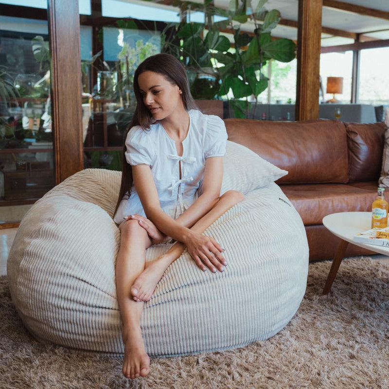 Woman sitting on a bean bag chair in a living room with a brown leather sofa and plants in the background.