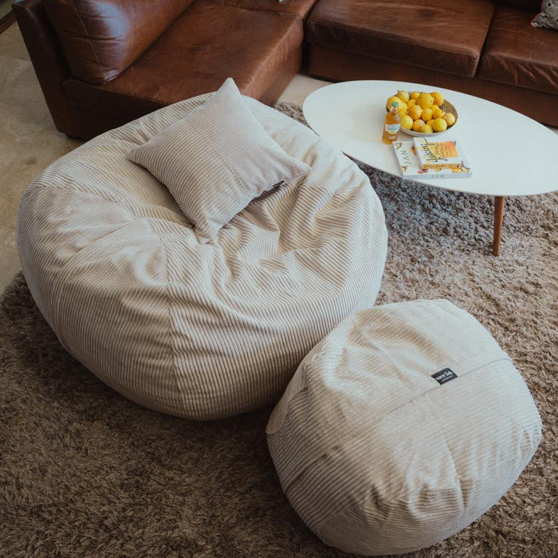 Beige bean bag chair and ottoman in a living room setting with a coffee table and fruit bowl.