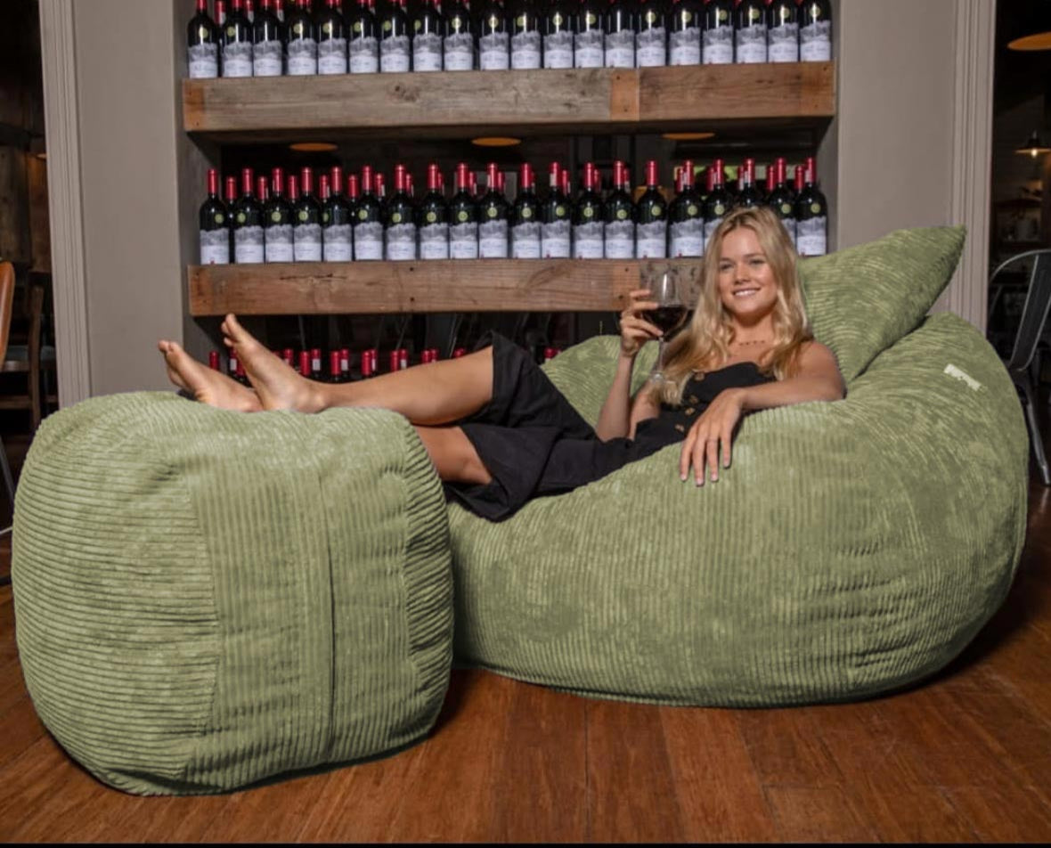 Woman sitting on a green bean bag chair holding a glass of wine with a wine rack in the background.
