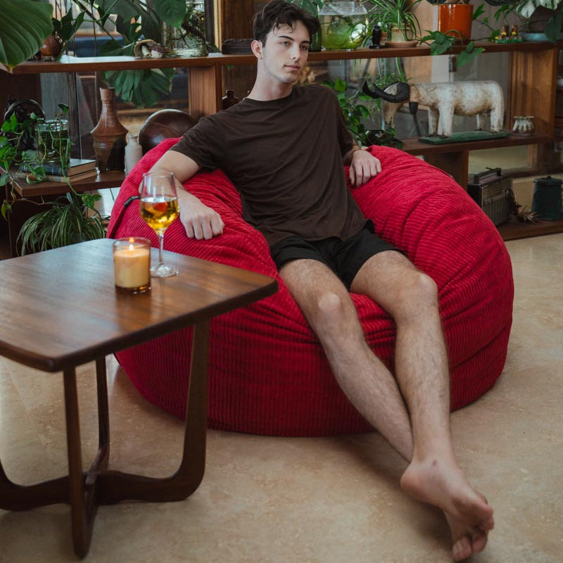 Man sitting on a red bean bag chair in a cozy room with plants and a candle.