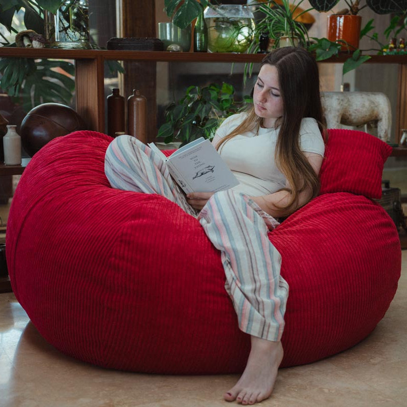 Woman reading a book on a large red bean bag chair in a cozy room with plants.