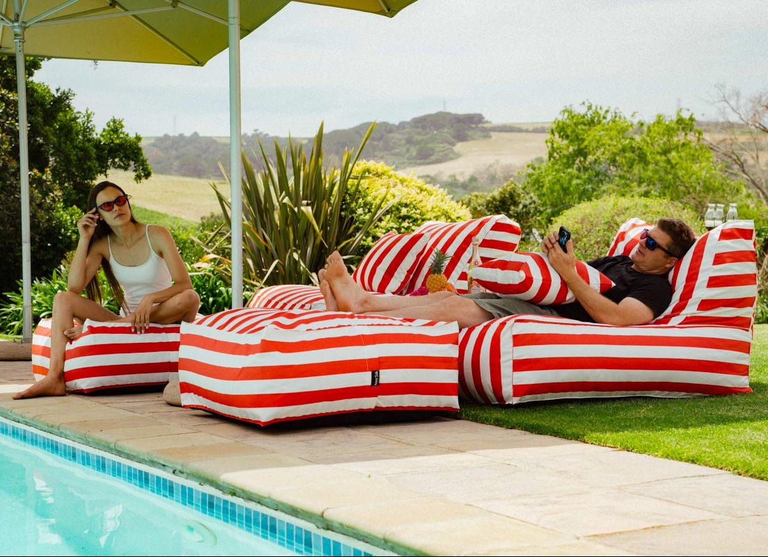 Two people relaxing on red and white striped lounge chairs by a pool with greenery in the background.