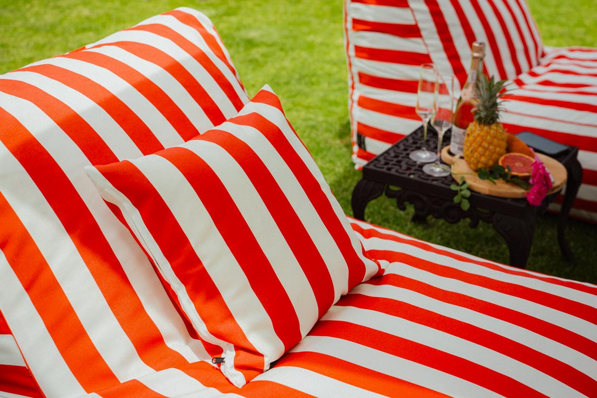 Red and white striped outdoor furniture with a small table holding drinks and fruit on a grassy background.