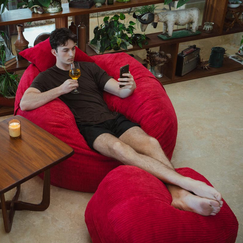 Man lounging on a red bean bag chair holding a phone and glass, with a small table and plants in the room.