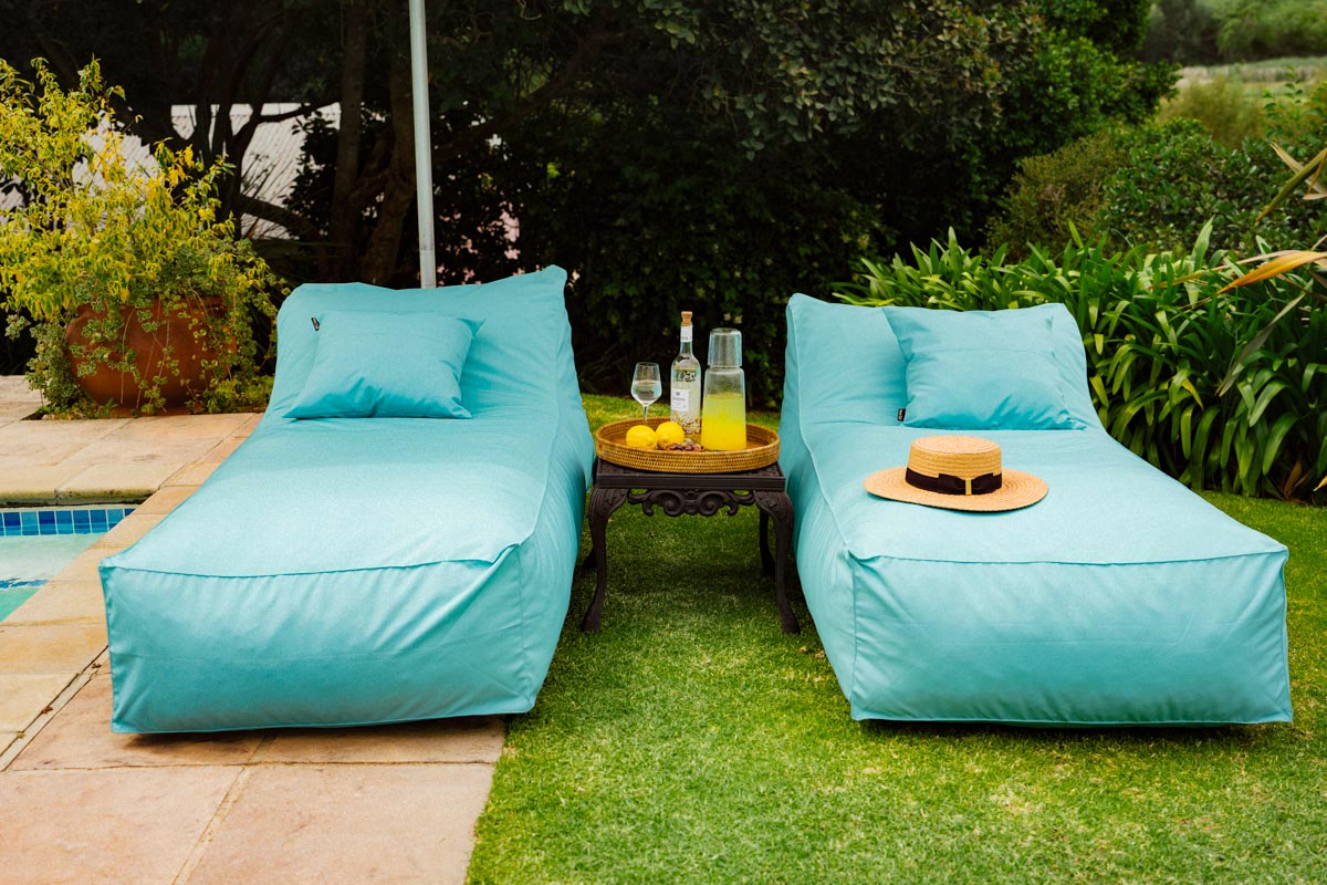 Two turquoise lounge chairs by a pool with a small table holding drinks and a hat.