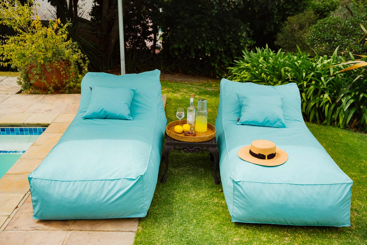 Two turquoise lounge chairs by a pool with a small table holding drinks and a hat.